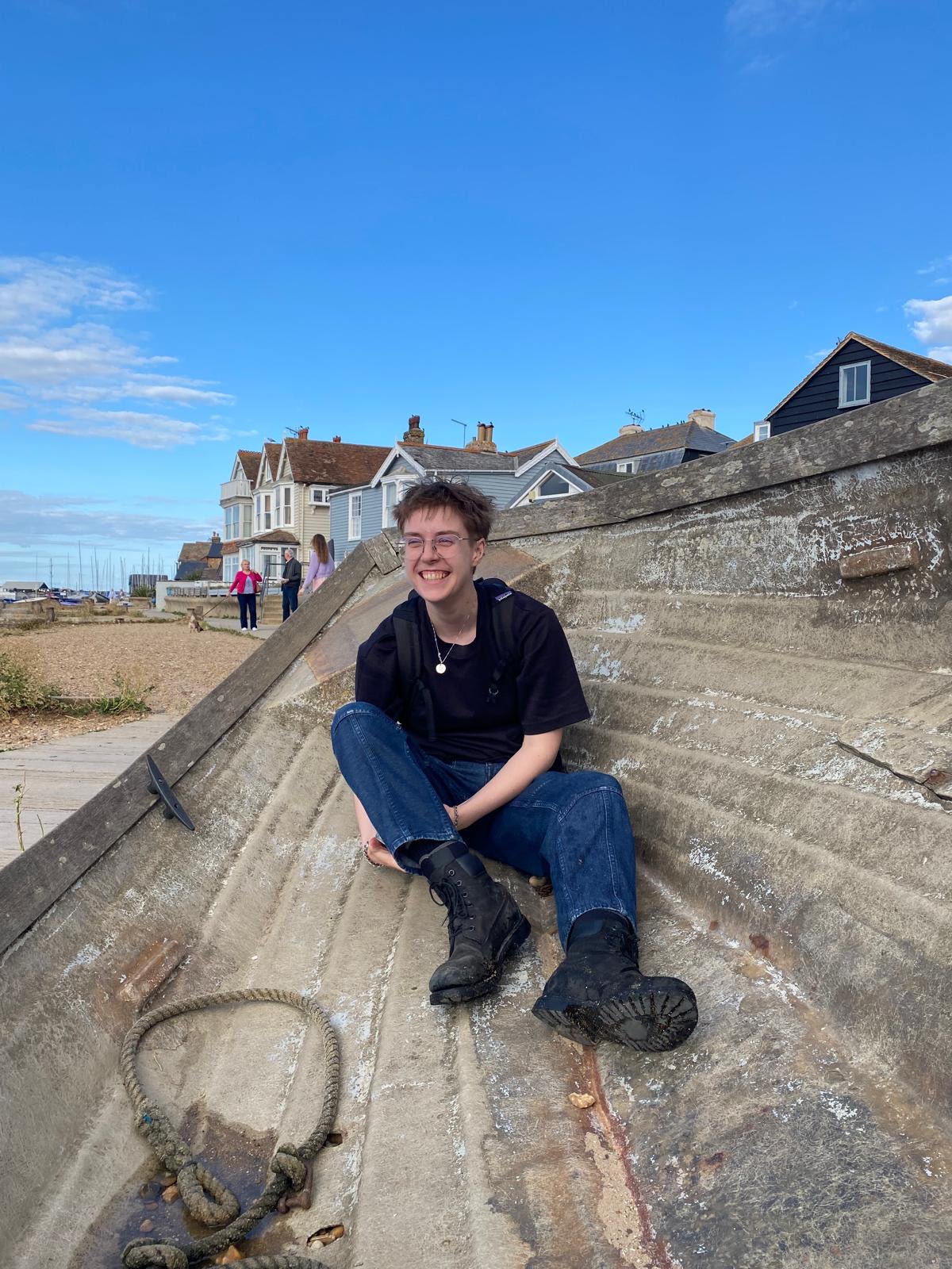 Young man wearing dark blue denim jeans, black boots and black top, with short wavy hair and glasses, sat in a wooden boat on a beach shore.
